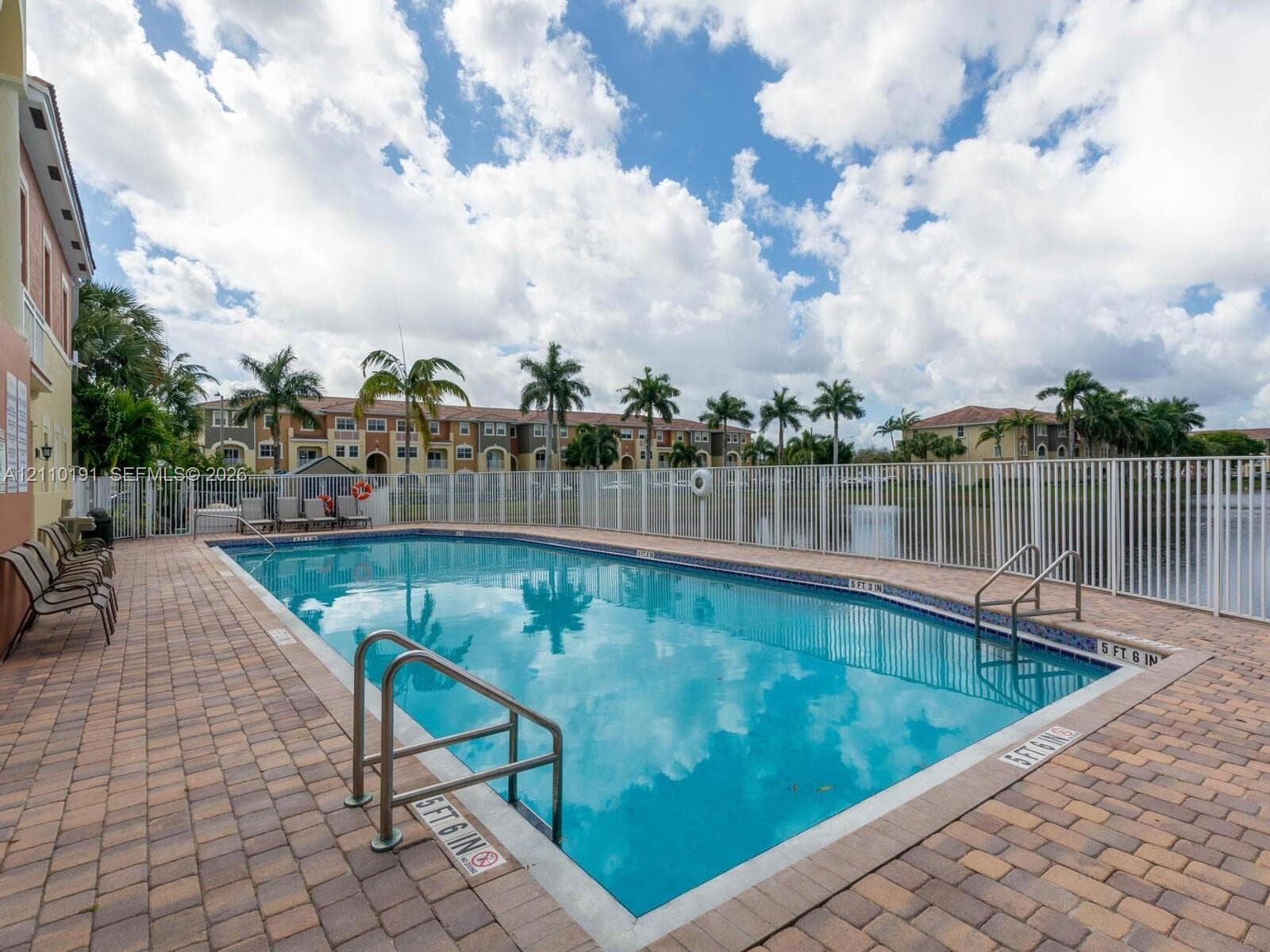 In-ground swimming pool with dual stainless steel entry railings, perimeter paver decking, and white metal safety fencing