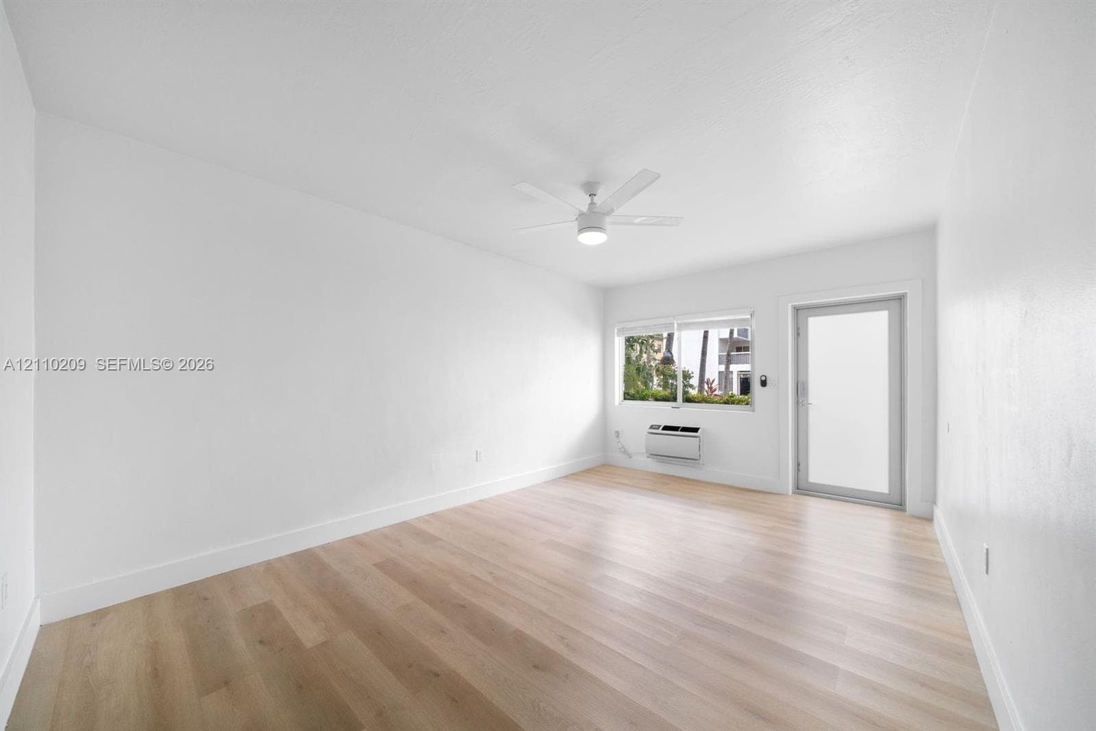 Room featuring wood-finish flooring, a white ceiling fan with integrated lighting, a wall-mounted air conditioning unit, and a glass-paneled exterior door