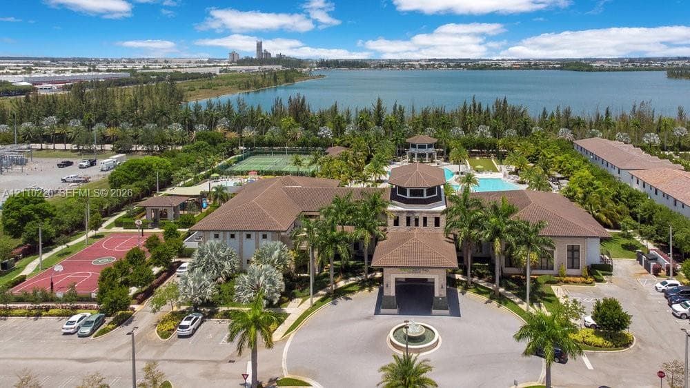 Aerial perspective of a multi-structure facility featuring a central fountain in a paved roundabout, a basketball court, tennis courts, and a swimming pool, situated on a lakefront lot