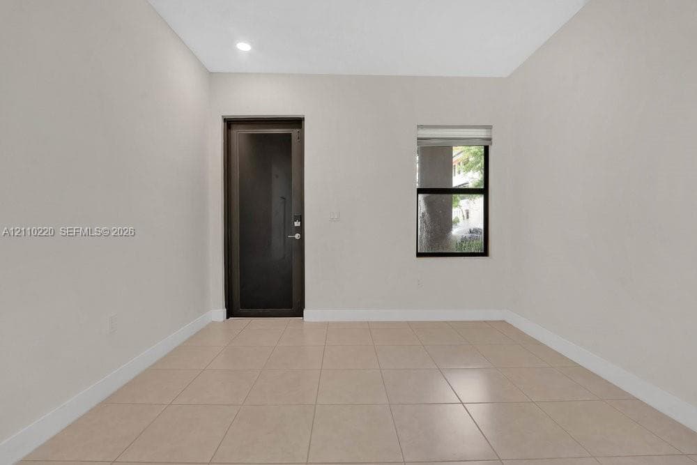 Neutral-toned interior room featuring large-format ceramic tile flooring, a dark-frame glass-panel door, a single-pane window with a roller shade, and recessed ceiling lighting