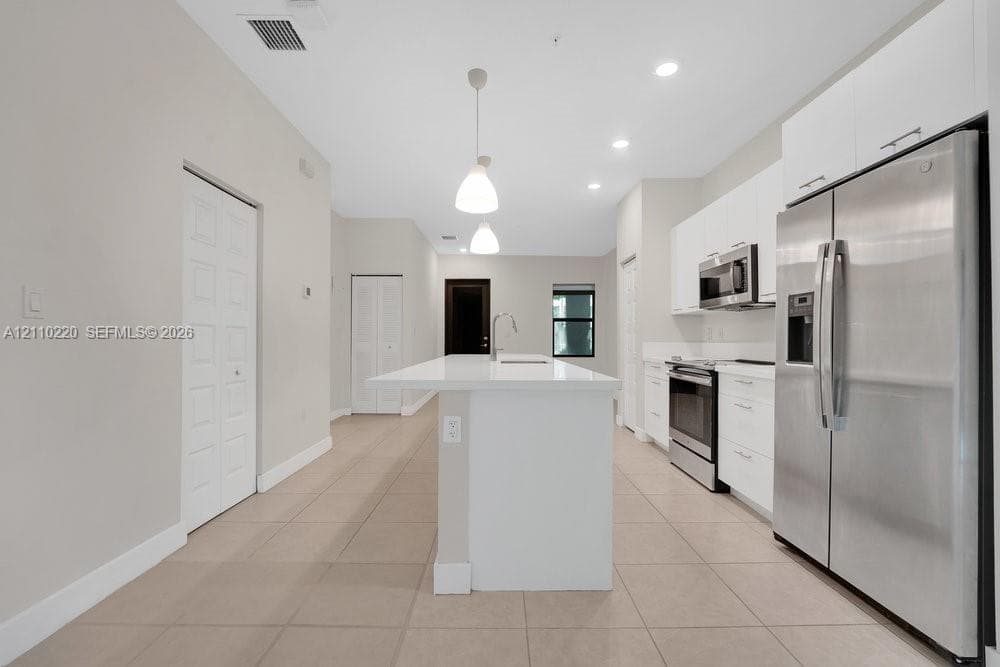 Kitchen featuring a central island with a solid-surface countertop, white flat-panel cabinetry, stainless steel appliances, recessed ceiling lighting, and large-format tile flooring