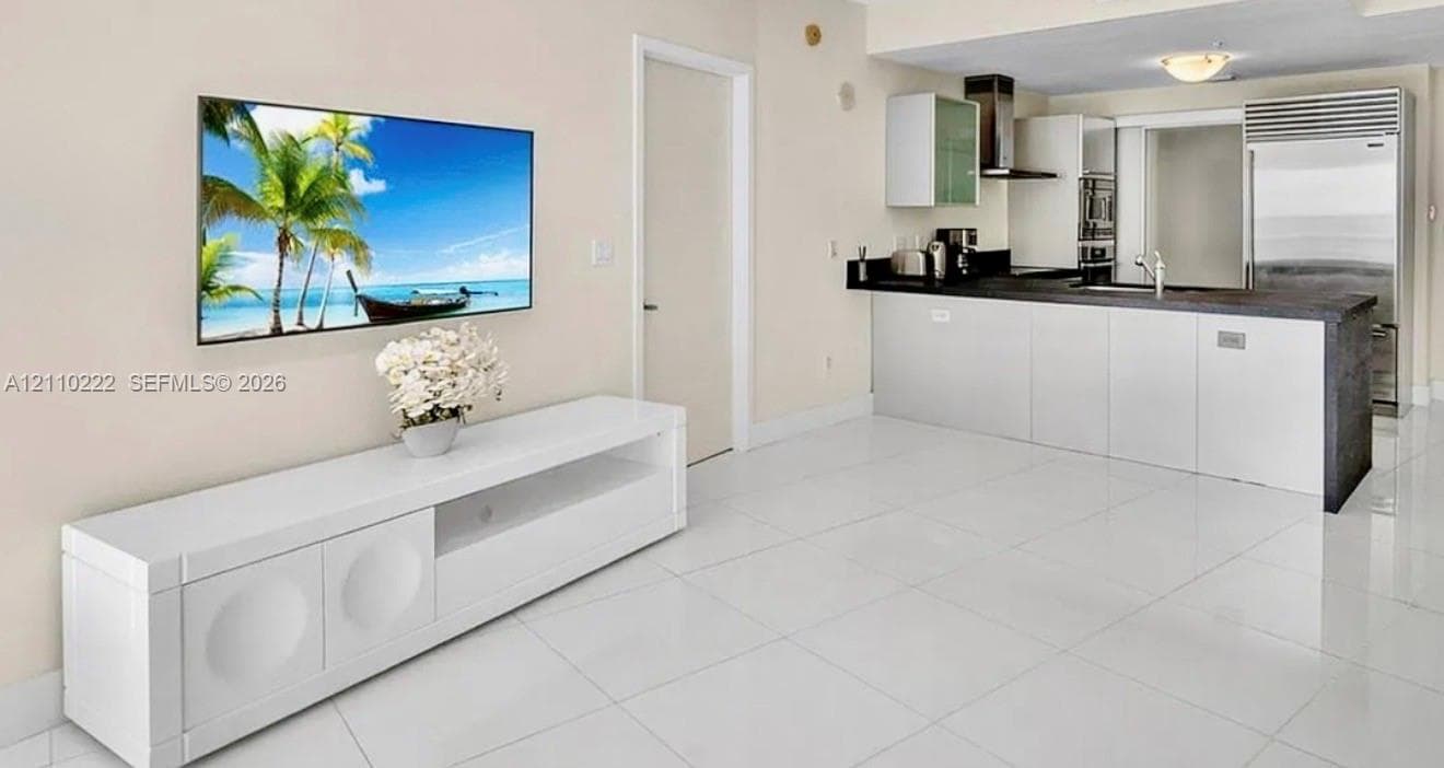 Open-concept kitchen featuring dark stone countertops, custom white cabinetry, stainless steel appliances, and polished white tile flooring