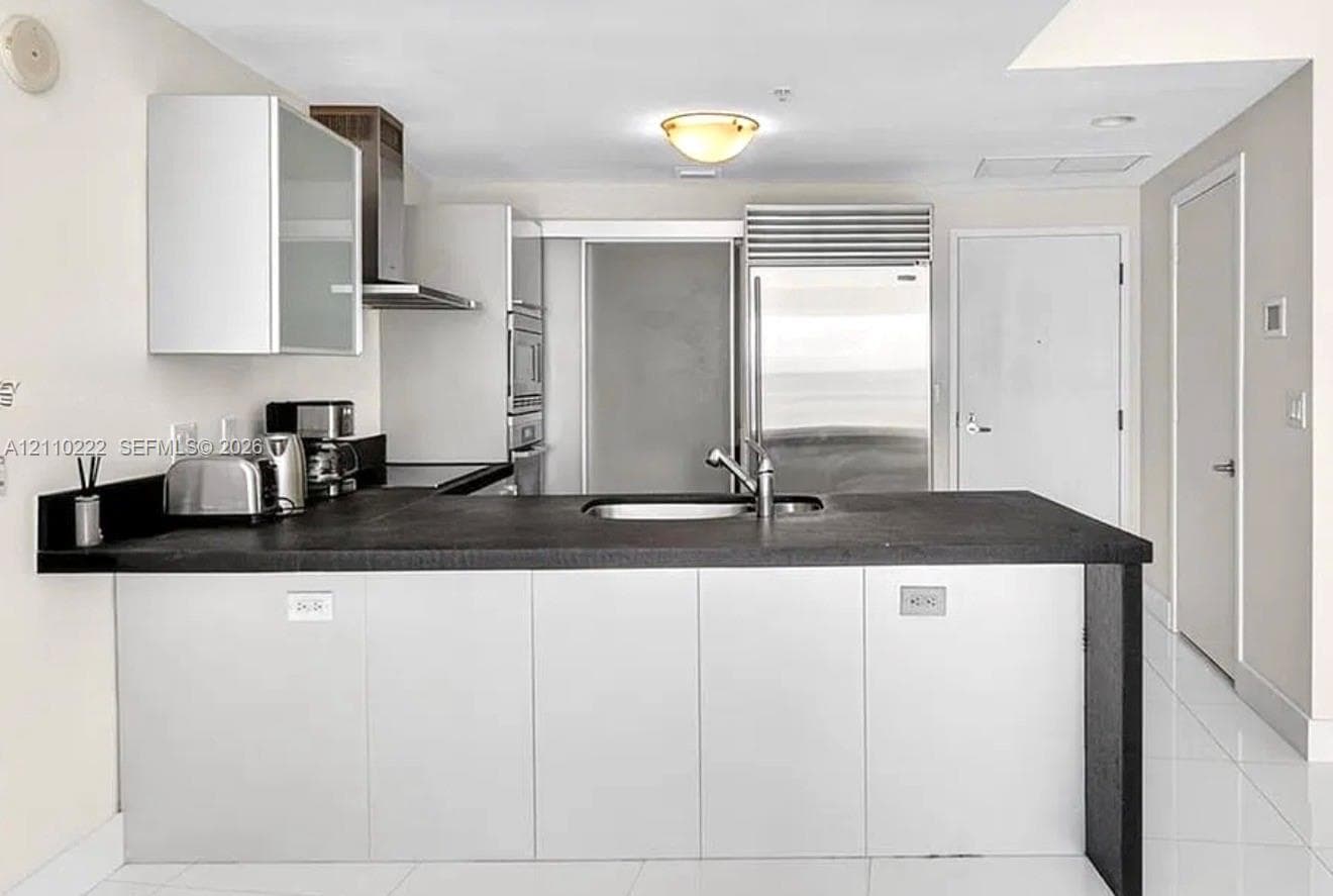 Kitchen featuring flat-panel cabinetry, a stainless steel refrigerator, stone-surfaced island with an integrated sink, and stainless steel range hood