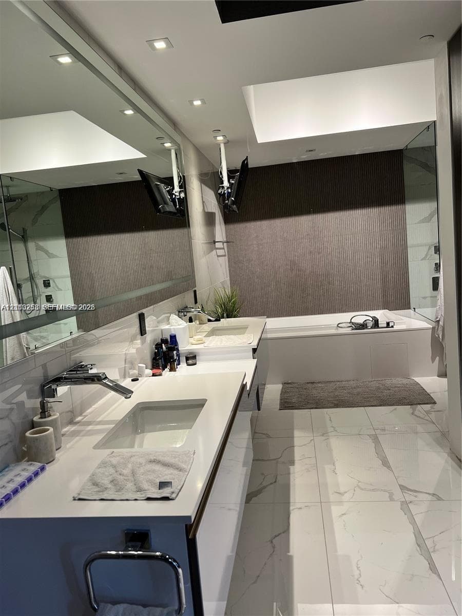 Modern bathroom featuring a double vanity with integrated rectangular sinks, white countertop, full-width mirror, recessed ceiling lighting, and a deep soaking tub with textured wall surround