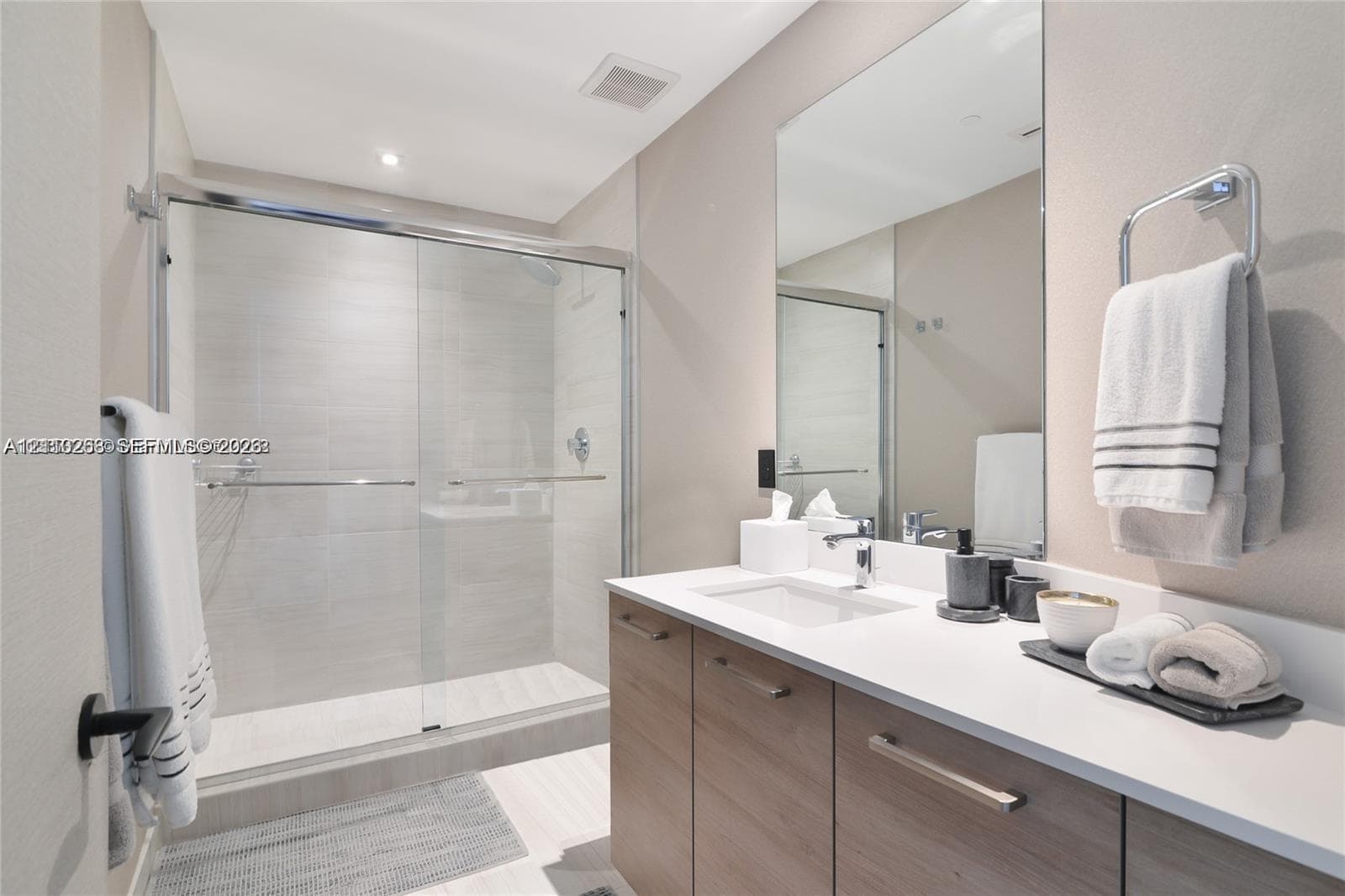 Bathroom featuring a vanity with light-tone wood-finish cabinetry, a solid-surface countertop with an integrated sink, a frameless glass shower enclosure with tiled walls, and a large wall-mounted mirror