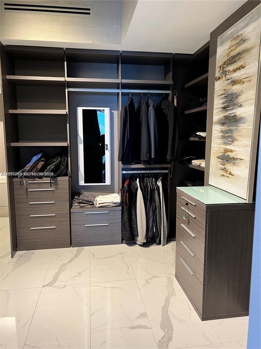 Custom closet system featuring dark wood-finish cabinetry, extensive built-in shelving, integrated dresser drawers with brushed metal pulls, and polished stone-finish flooring