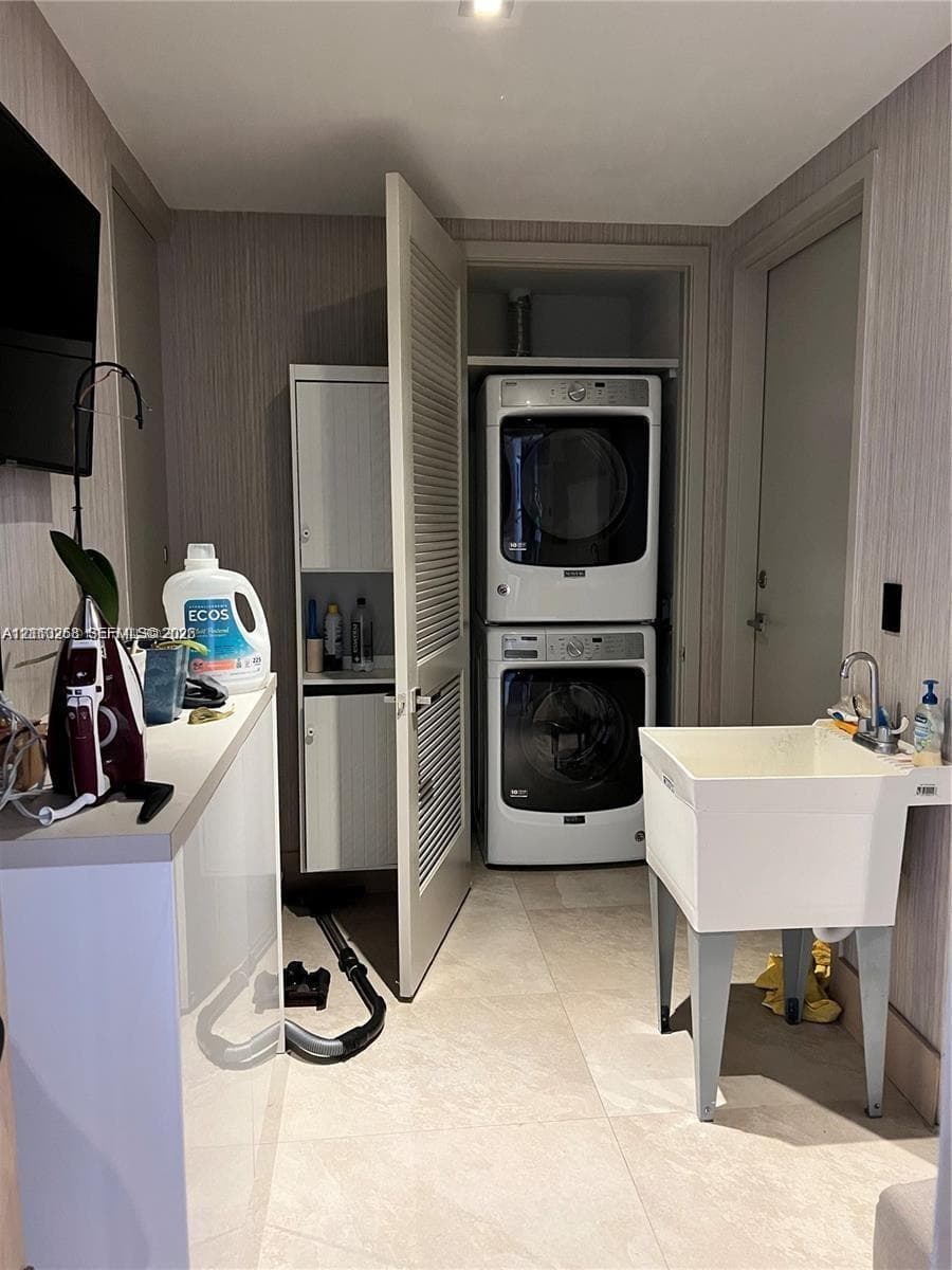 Laundry area featuring stacked white washer and dryer, built-in white cabinetry, louvered closet door, freestanding utility sink with chrome faucet, and tile flooring