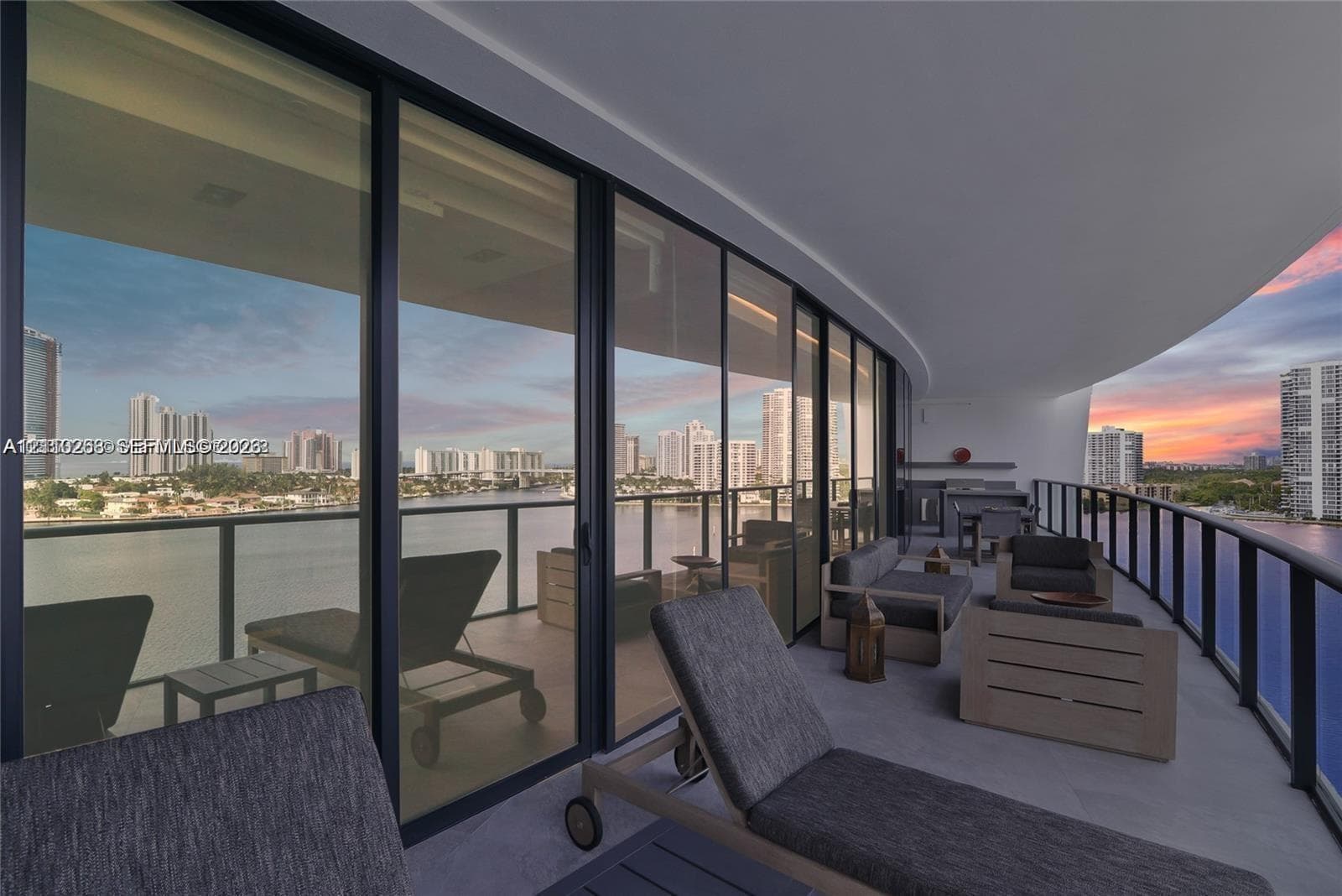 Expansive covered balcony featuring floor-to-ceiling glass doors, a built-in outdoor kitchen, and dark metal railings, overlooking a waterfront skyline