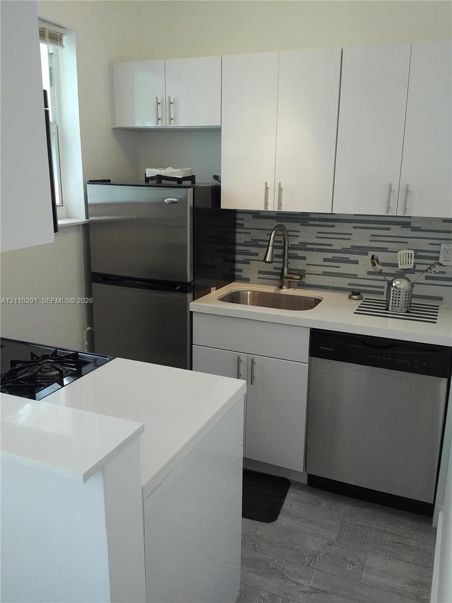 Kitchen featuring white flat-panel cabinetry, stone-composite countertops, stainless steel appliances, multi-tonal linear tile backsplash, and wood-finish flooring