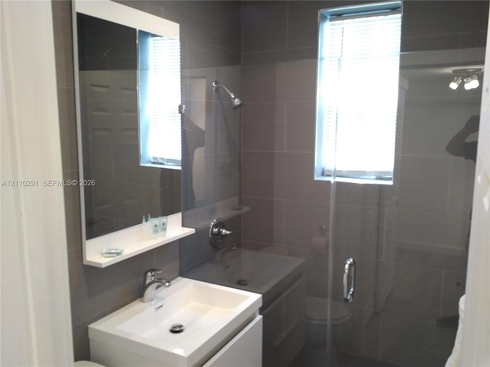 Modern floating vanity with a rectangular sink, wall-mounted mirror with integrated shelf, glass-enclosed shower, polished chrome fixtures, and grey ceramic wall tiling