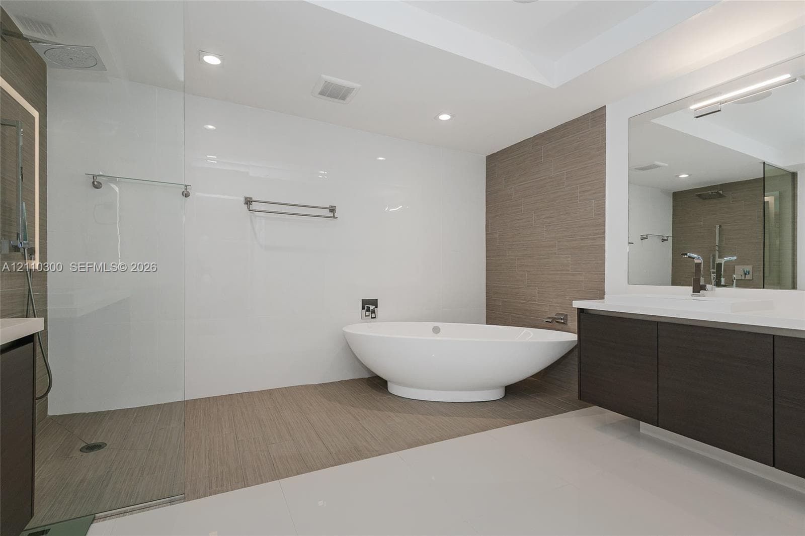 Master Bathroom Floating vanity with dark wood-finish cabinetry, integrated LED-backlit mirror, walk-in glass shower, wall-mounted shower hardware, and freestanding soaking tub