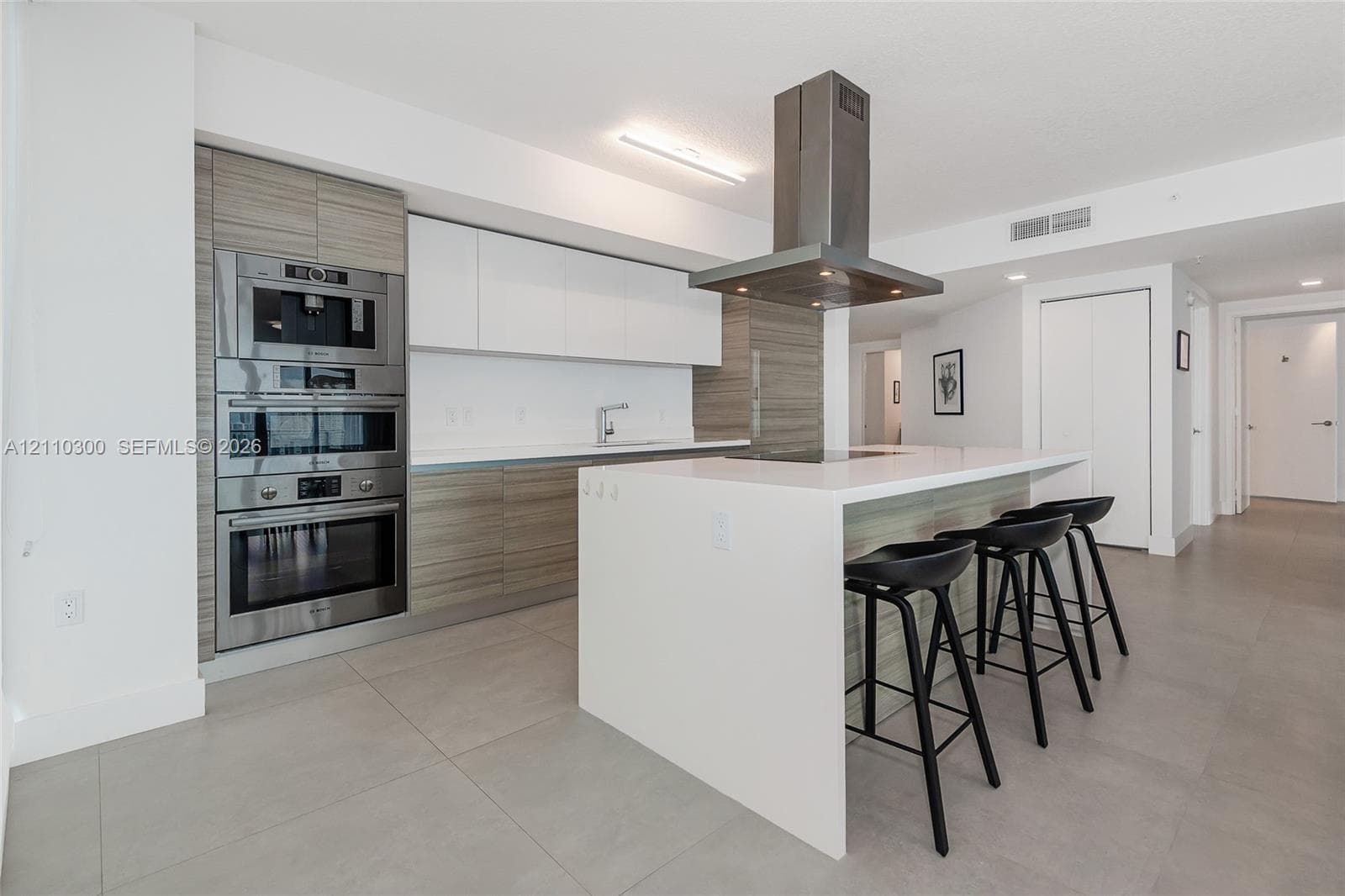 Modern kitchen featuring an island with a waterfall-edge countertop, panel-ready refrigerator, stainless steel ceiling-mounted range hood, built-in double wall ovens, and two-tone flat-panel cabinetry
