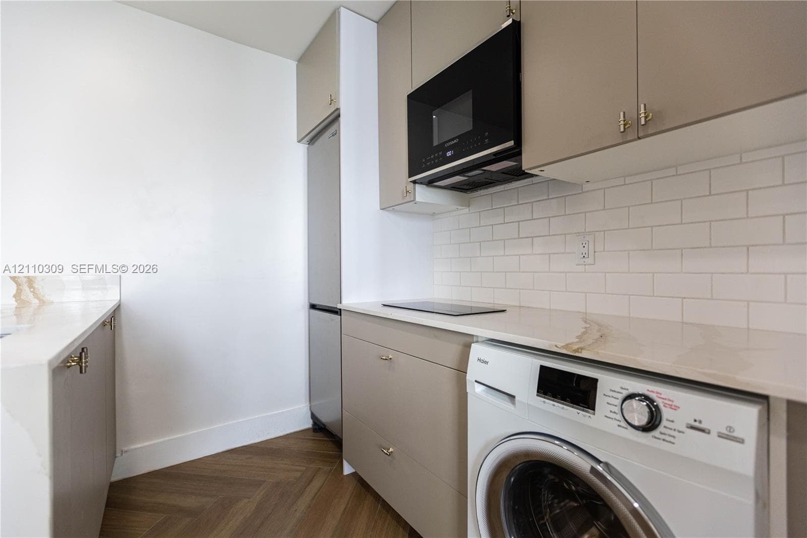 Contemporary kitchen featuring neutral flat-panel cabinetry, white subway tile backsplash, stone-finish countertops with integrated cooktop, stainless steel appliances, and herringbone-patterned wood-finish flooring
