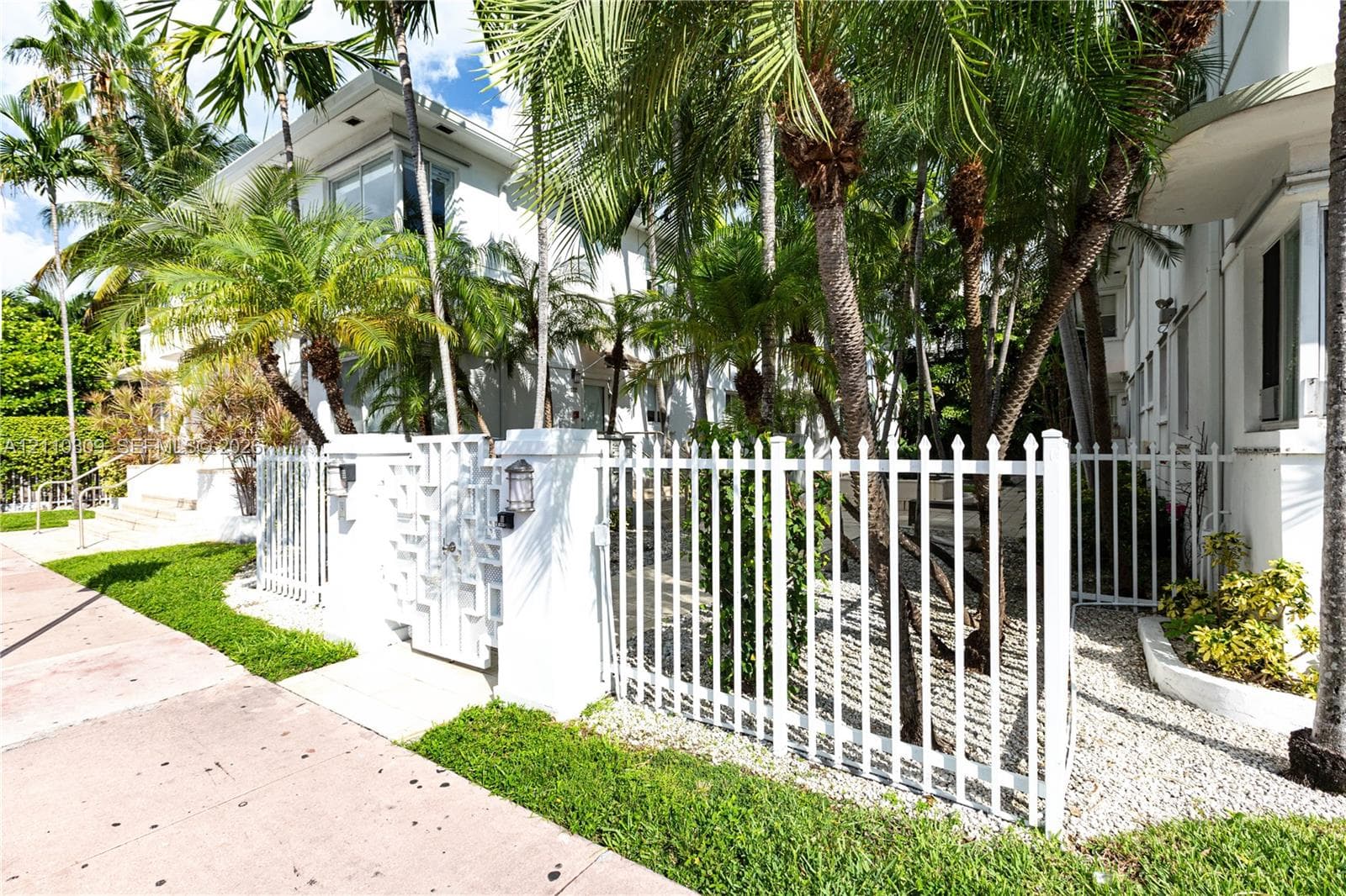White exterior building facade featuring a gated entry, perimeter fencing, and mature palm trees