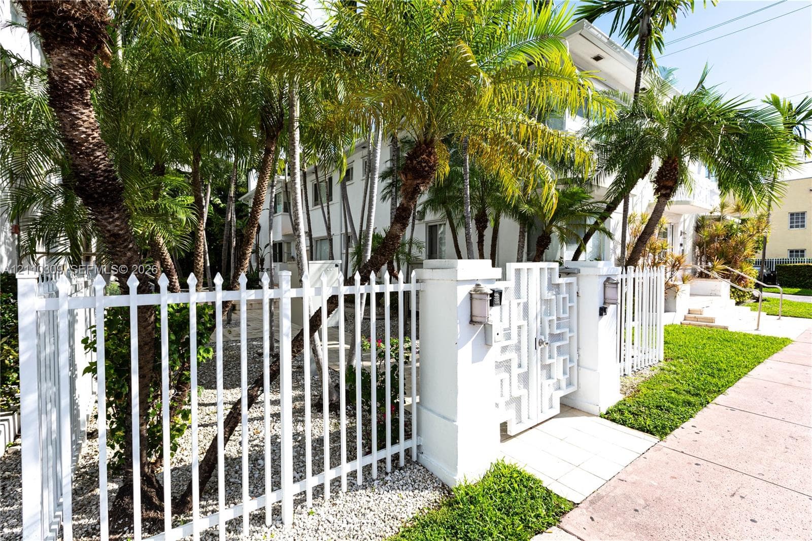 White metal perimeter fencing, decorative geometric gate, stucco exterior facade, and mature palm trees