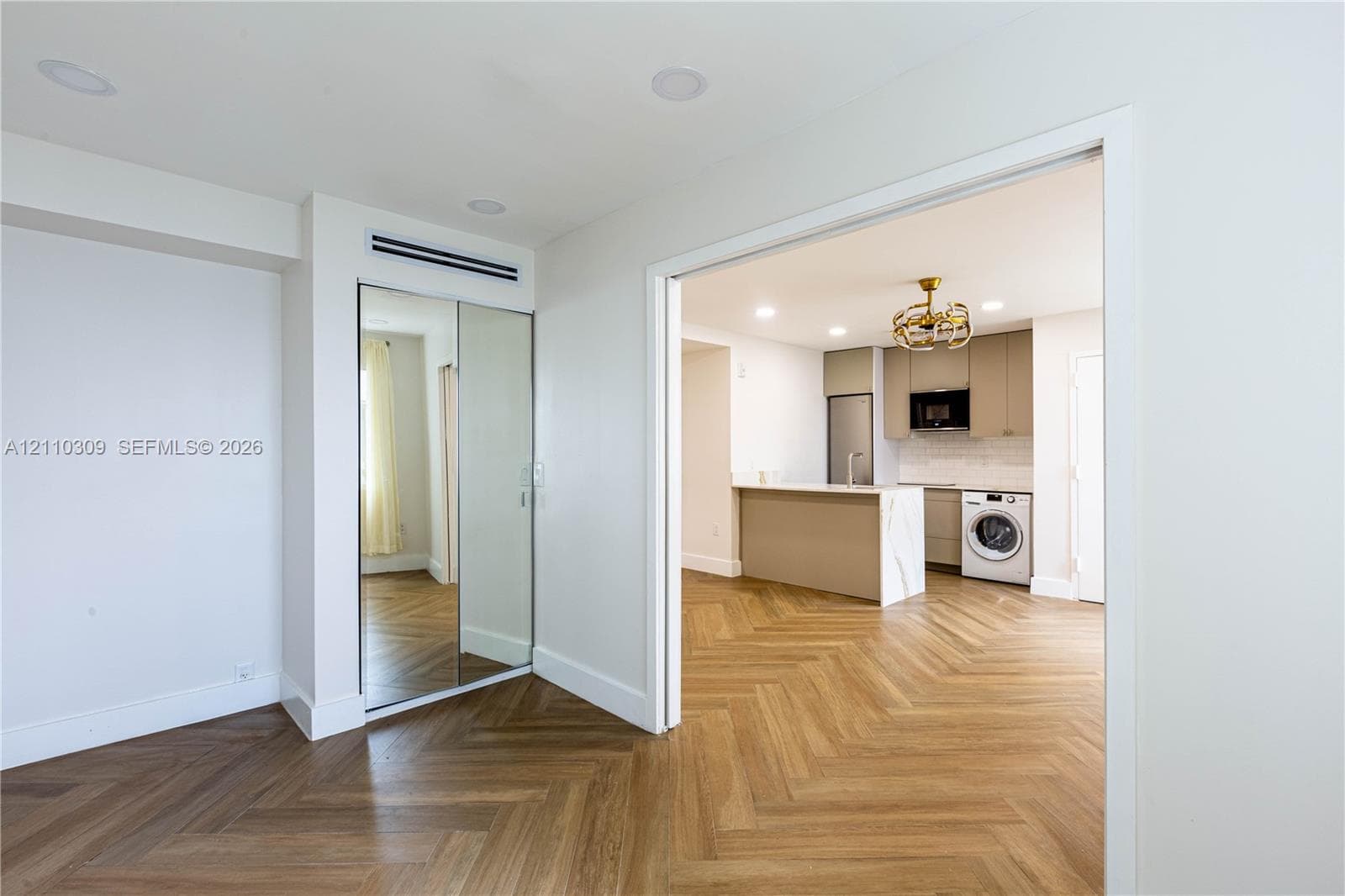 Herringbone wood-finish flooring, mirrored sliding-door closet, recessed ceiling lighting, kitchen island with stone-finish waterfall countertop, and built-in laundry appliance
