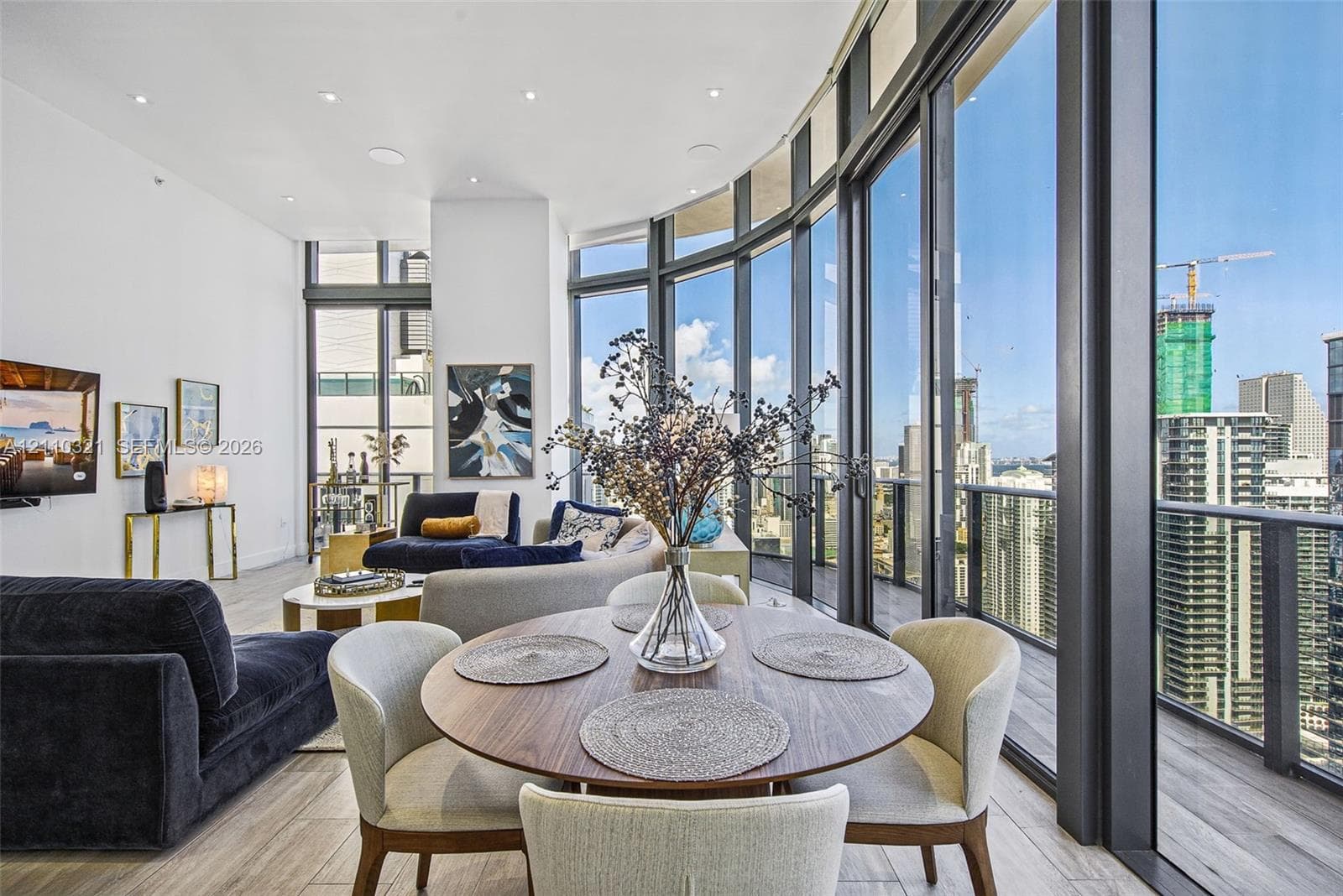 Curved floor-to-ceiling windows, wrap-around private balcony with glass railing, wood-finish flooring, recessed lighting, and urban skyline panorama
