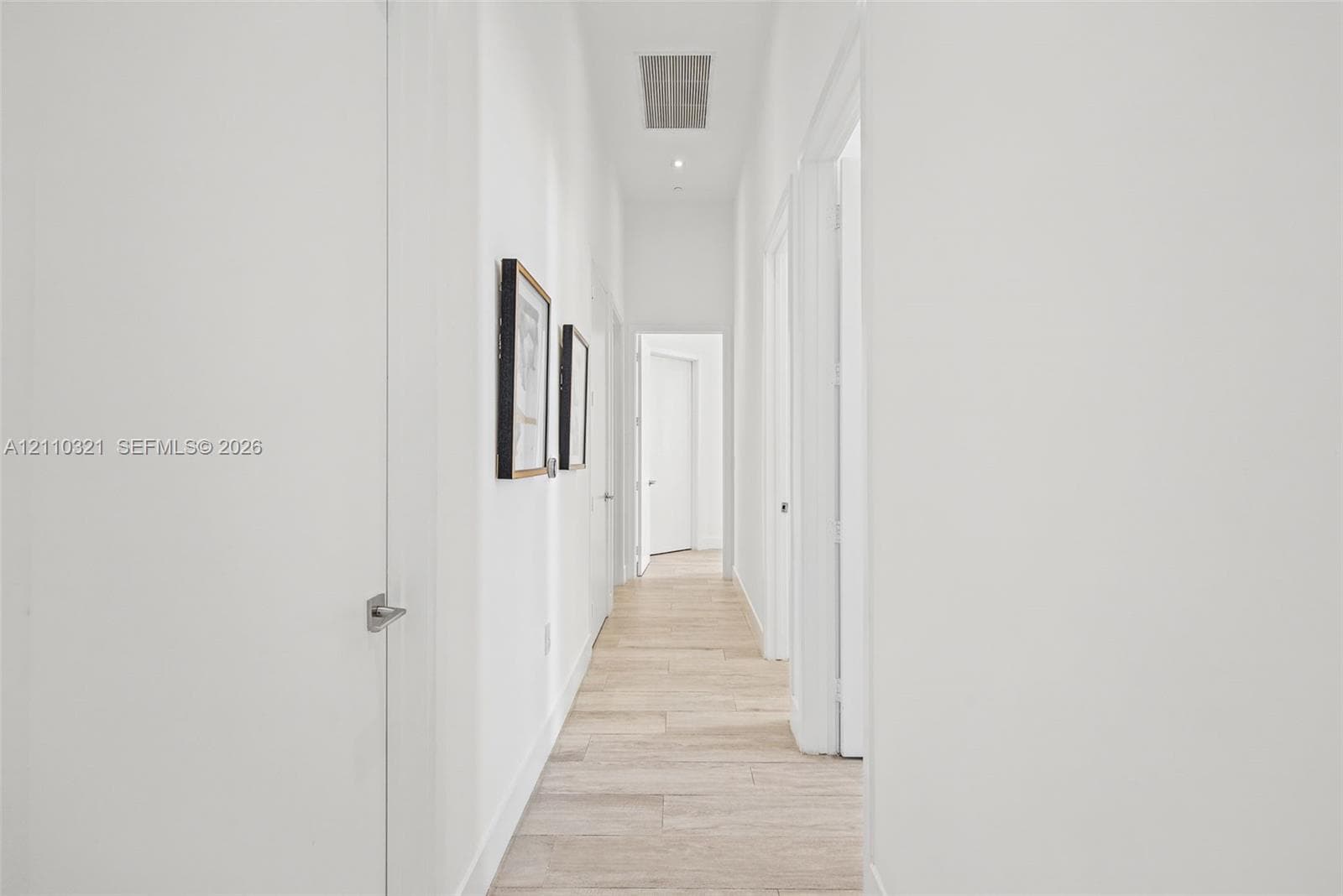 Hallway featuring light-toned wood-finish flooring, crisp white wall finishes, and multiple interior swing doors with satin nickel hardware