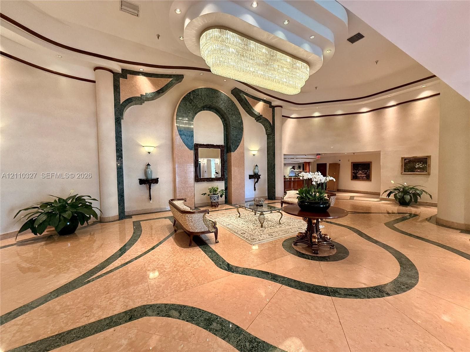 Grand foyer featuring polished stone flooring with intricate inlaid stone accents, a multi-tiered recessed ceiling, a large-scale crystal chandelier, and architectural wall detailing with stone arches