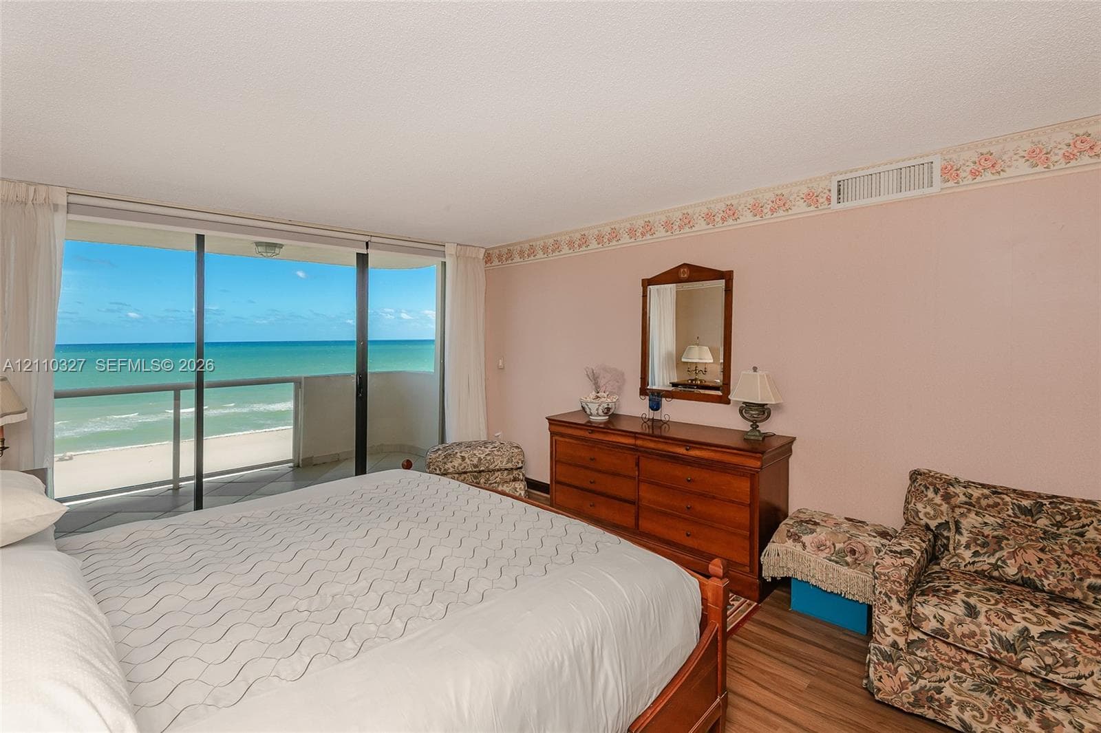 Floor-to-ceiling sliding glass doors opening to a private balcony, wood-finish flooring, and ocean backdrop
