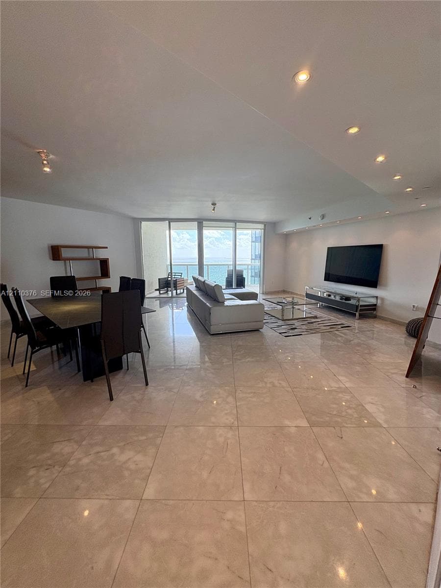 Open-concept living area featuring large-format tile flooring, recessed ceiling lighting, and floor-to-ceiling glass sliding doors leading to a balcony