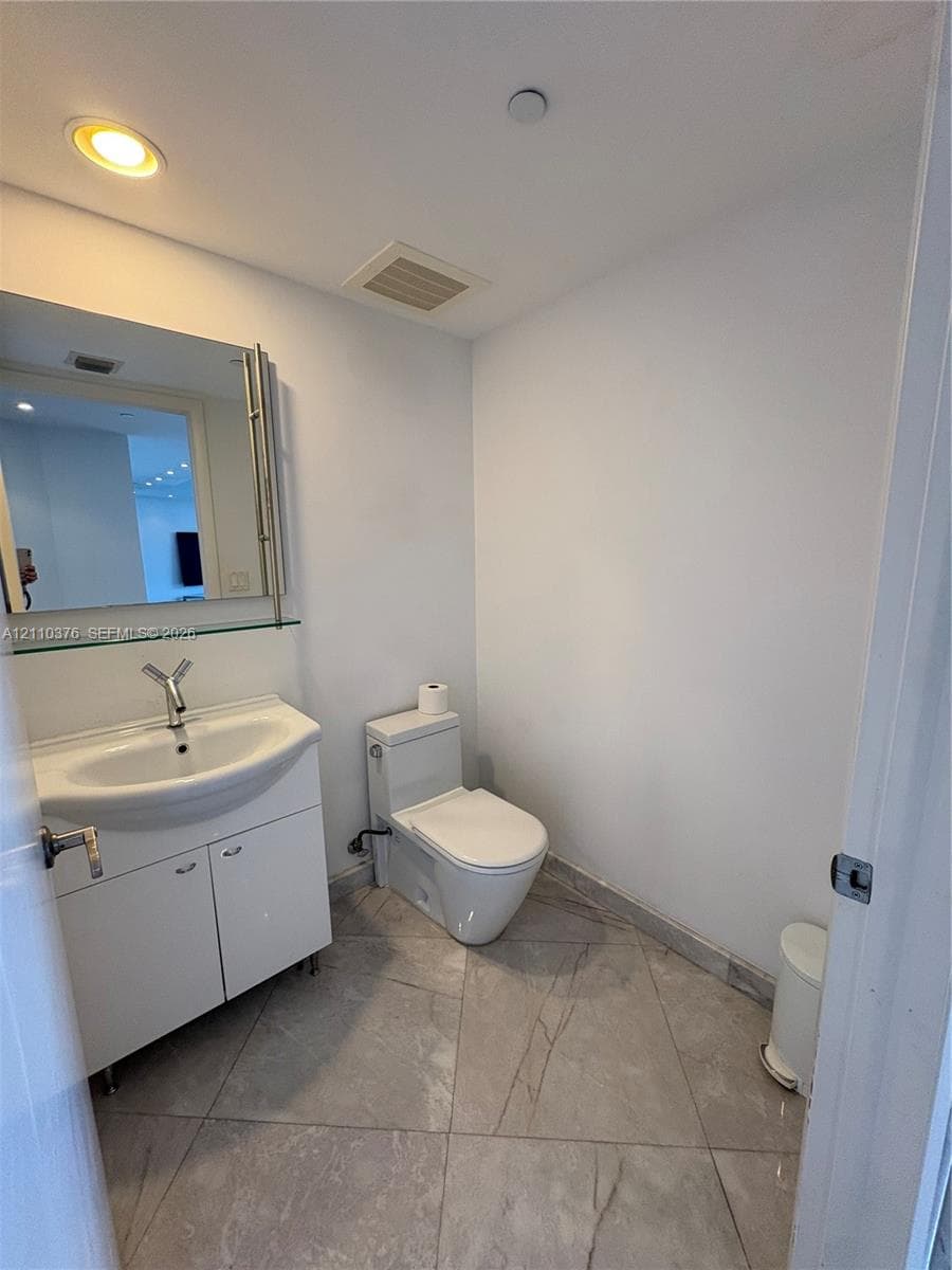 White cabinetry vanity with integrated sink, polished metal faucet, wall-mounted mirror with glass shelving, and large-format tile flooring