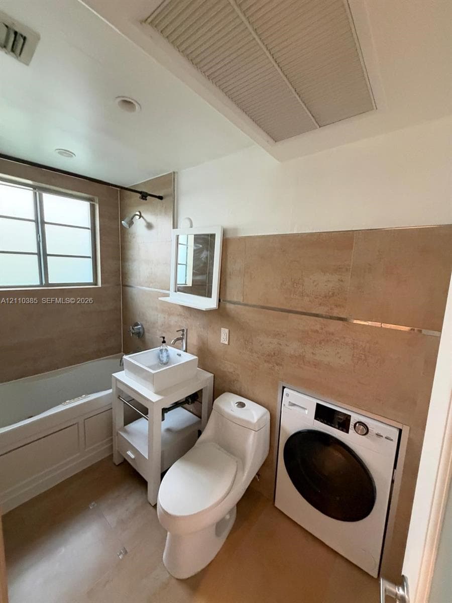 Bathroom featuring full-height neutral wall tiling, an integrated front-loading washer, a white pedestal vanity with a rectangular vessel sink, and a combination soaking tub and shower with a metal trim finish
