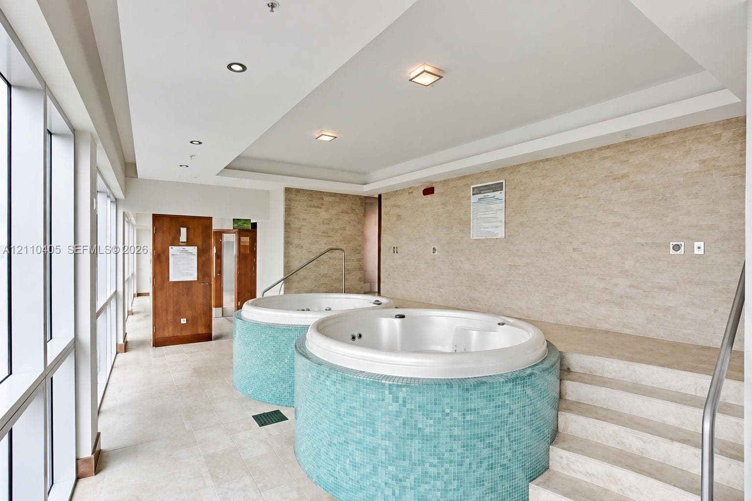 Spa facility featuring dual circular jetted tubs with turquoise tile surrounds, natural stone wall cladding, recessed lighting, and tiered stone steps with a metal handrail