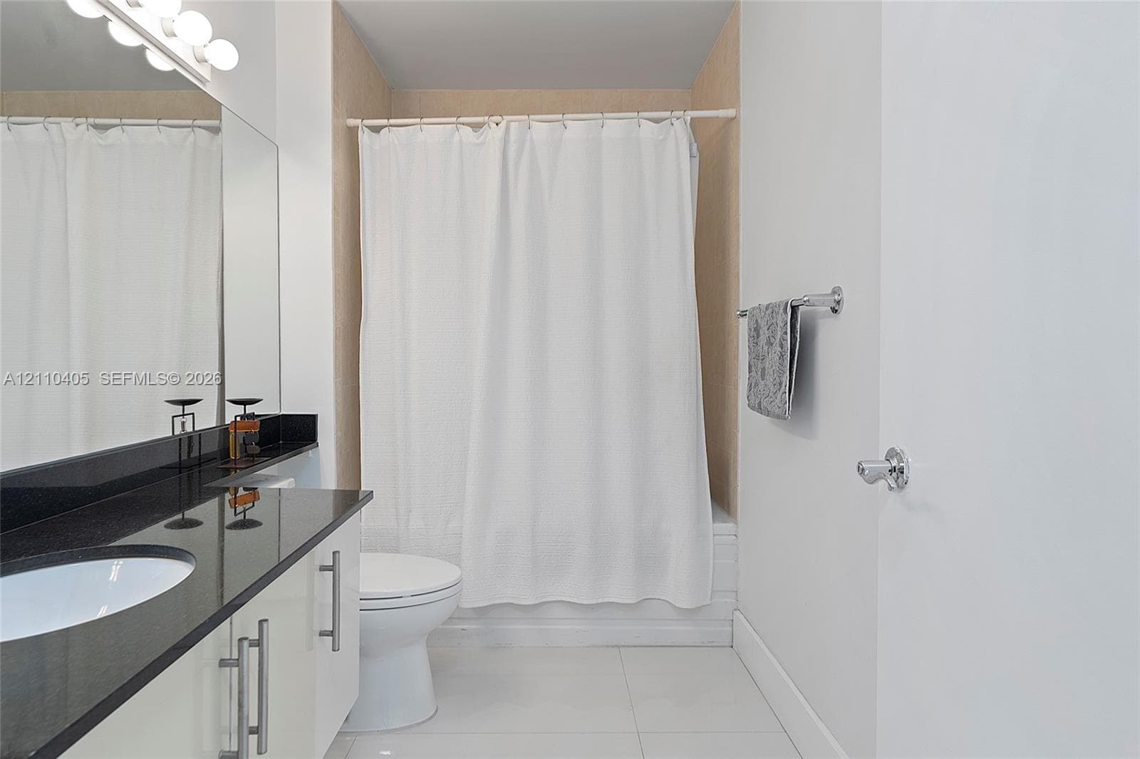 Bathroom featuring a vanity with dark stone countertop, integrated sink, polished metal hardware, full-width mirror with integrated lighting, and a bathtub with tile surround