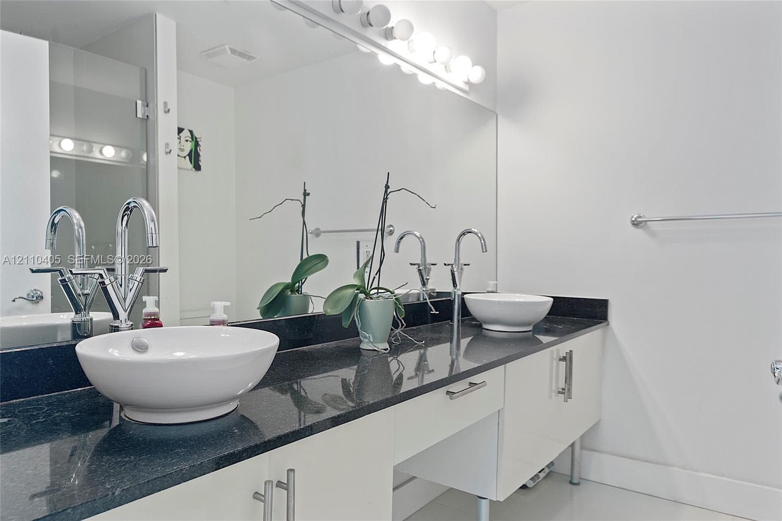 Dual vanity featuring a dark countertop, white high-gloss cabinetry, vessel sinks with polished chrome faucets, and a full-width mirror with integrated lighting