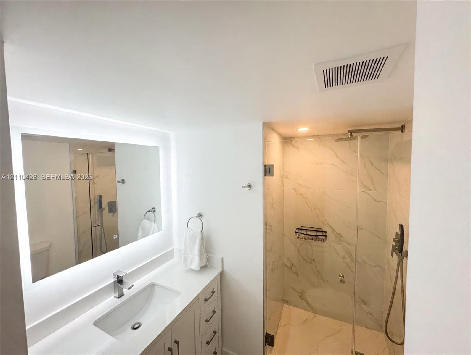 Bathroom featuring a white vanity with a solid-surface countertop, an integrated sink, an LED-backlit mirror, and a walk-in shower with large-format stone-patterned tile and a glass enclosure