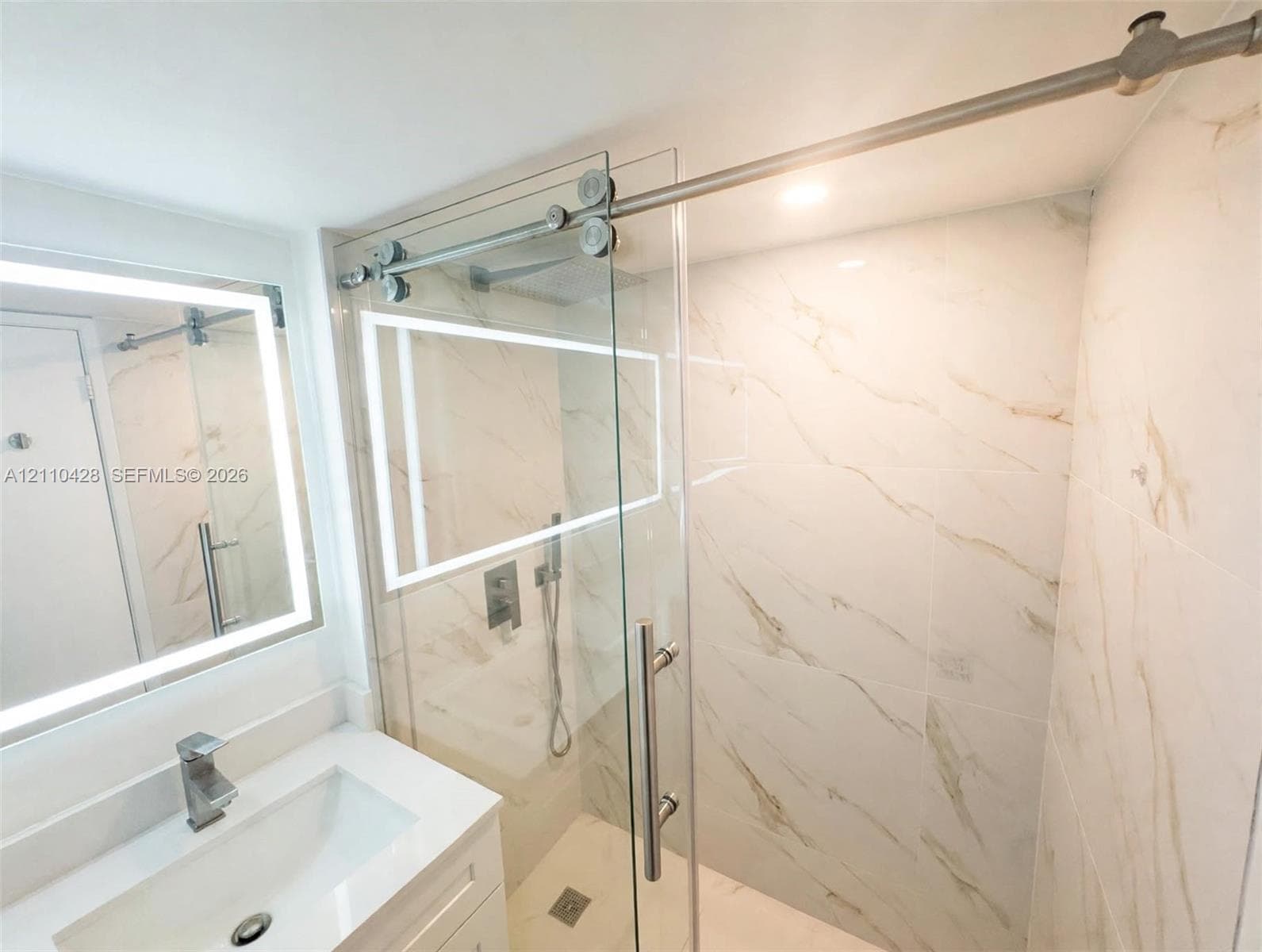 Frameless glass shower enclosure with stainless steel sliding hardware, full-height stone-finish wall tile, illuminated vanity mirror, and single-lever faucet