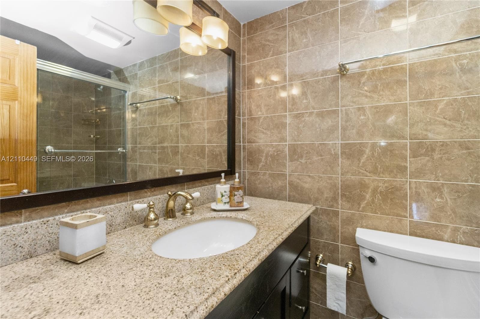 Bathroom featuring a vanity with a stone-surfaced countertop, an undermount sink with brass-finish hardware, floor-to-ceiling tile work, and a framed wall mirror with multi-bulb light fixture