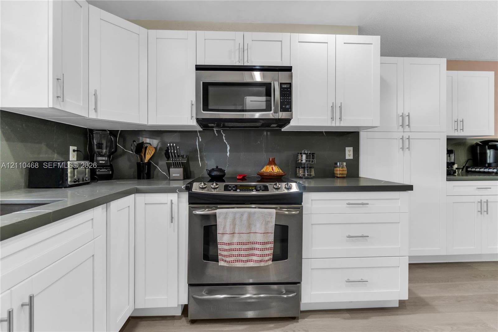 Kitchen featuring shaker-style white cabinetry, dark solid-surface countertops, full-height backsplash, stainless steel appliances, and wood-finish flooring