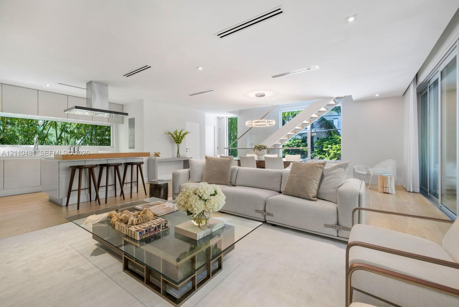 Open-concept interior featuring light wood-finish flooring, linear recessed lighting, a modern floating staircase with glass railings, large horizontal windows, and a stainless steel kitchen ventilation hood