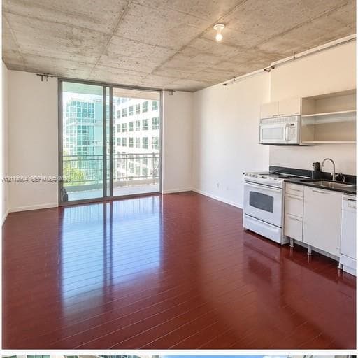 Exposed concrete ceiling, wood-finish flooring, sliding glass door with balcony access, and kitchen featuring white cabinetry and built-in appliances