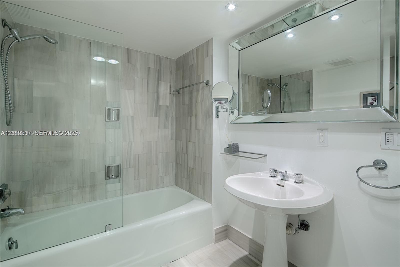 Bathroom featuring a white pedestal sink, frameless glass shower screen, tub with wall-mounted shower head, large beveled mirror, and stone-finish wall tile