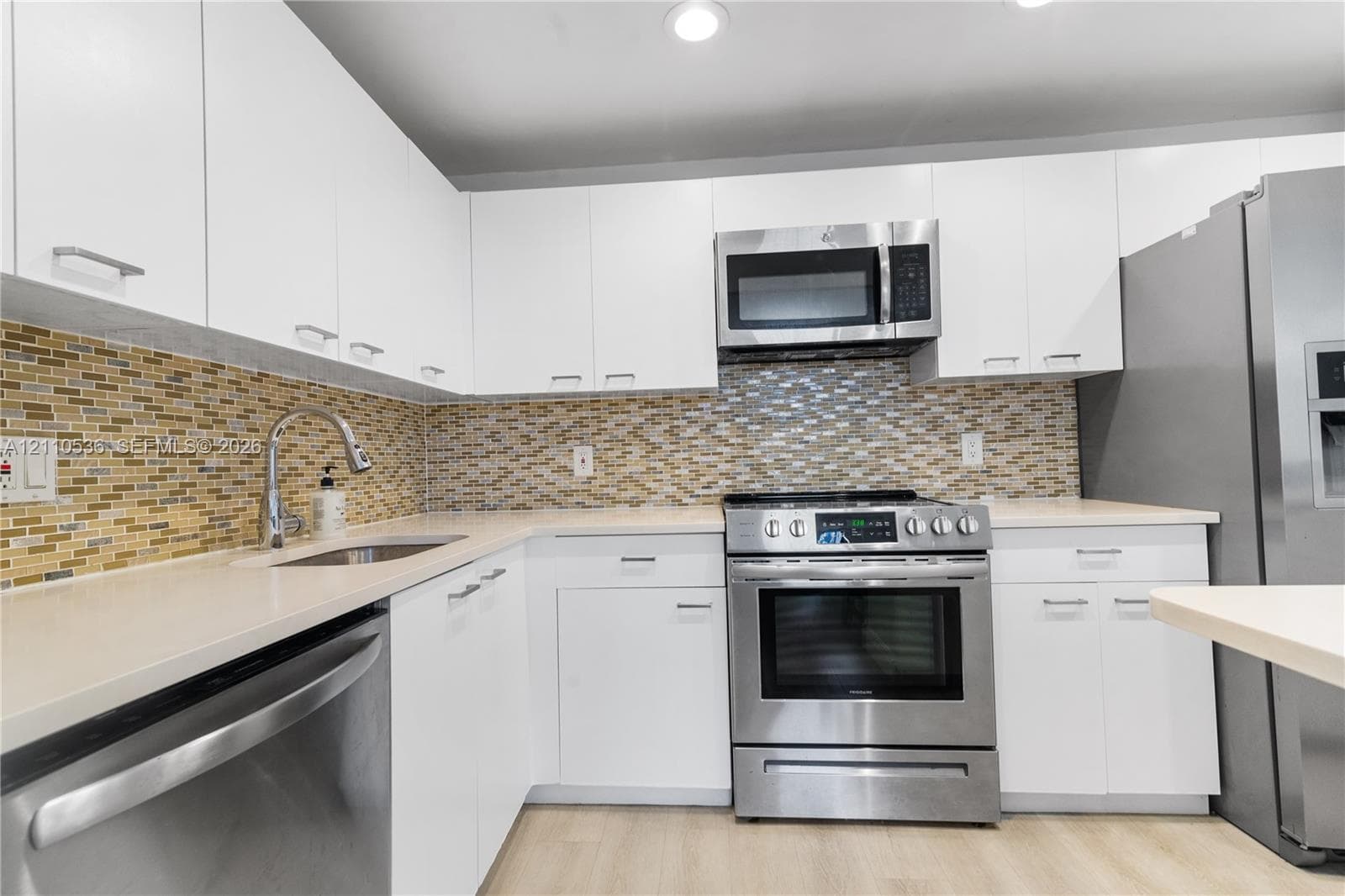 Kitchen featuring flat-panel white cabinetry, stainless steel appliances, multi-tonal mosaic tile backsplash, solid-surface countertops, and wood-finish flooring