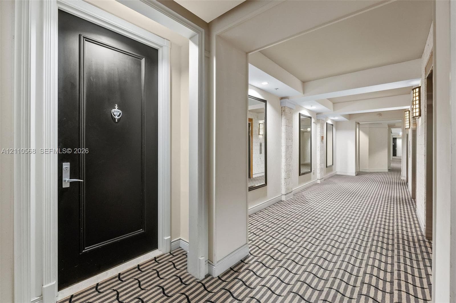 Black paneled entry door with chrome hardware, patterned wall-to-wall carpeting, recessed ceiling lighting, wall-mounted rectangular mirrors, and architectural pillars