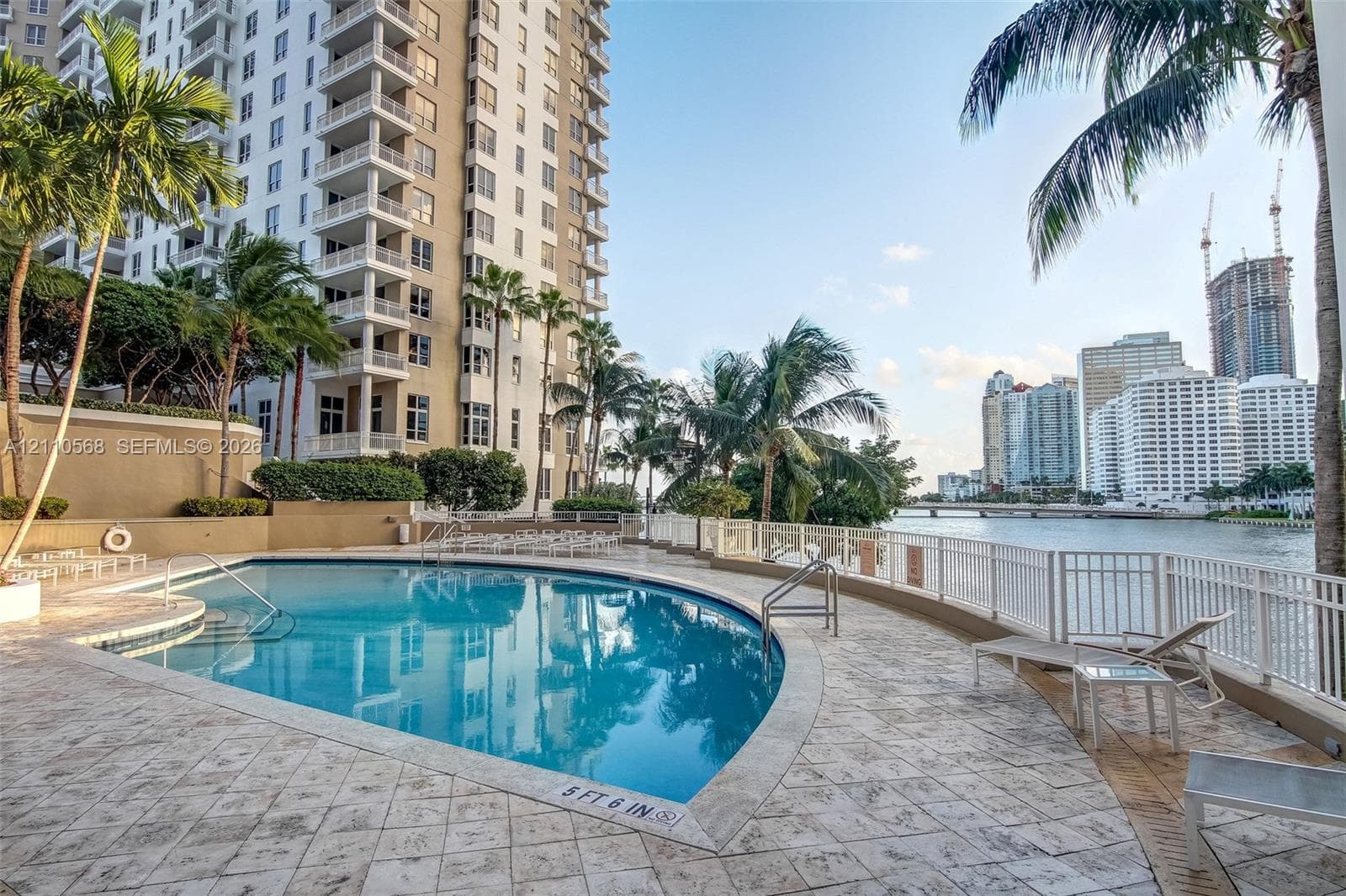 Waterfront swimming pool with a stone deck, perimeter safety fencing, and surrounding palm trees