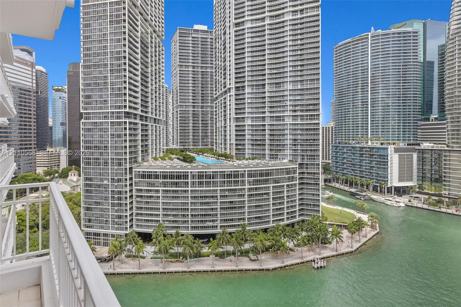 High-rise balcony with white metal railing overlooking a waterfront promenade, palm tree landscaping, and a multi-level complex featuring an elevated swimming pool