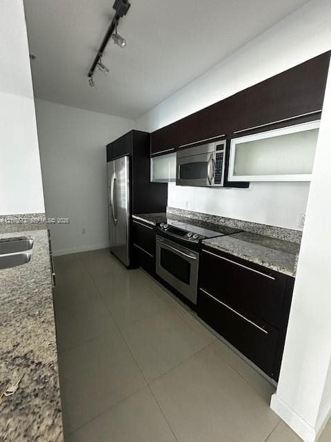 Kitchen featuring dark espresso cabinetry, stone countertops with matching backsplash, stainless steel appliances, frosted glass cabinet inserts, and tile flooring