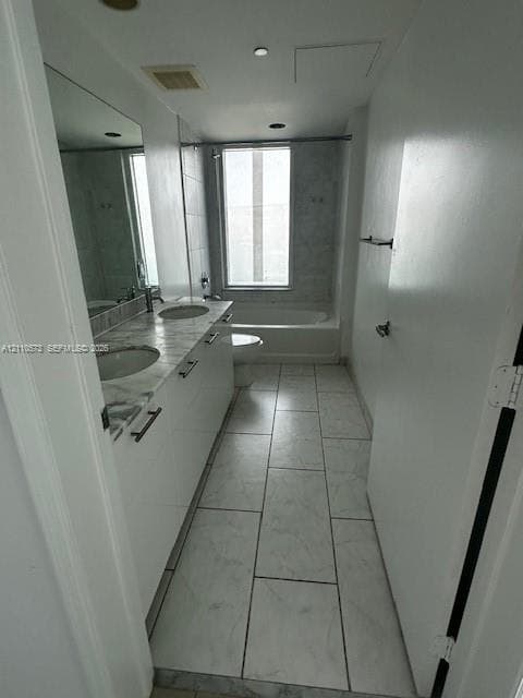 Dual-sink vanity with stone-finish countertop, white cabinetry with dark hardware, full-length mirror, large-format tile flooring, and a bathtub with tile surround