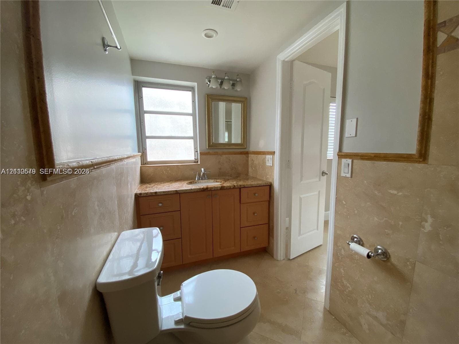 Bathroom featuring stone-tile wall wainscoting, wood-finish cabinetry, natural stone countertop, undermount sink, and wall-mounted vanity mirror