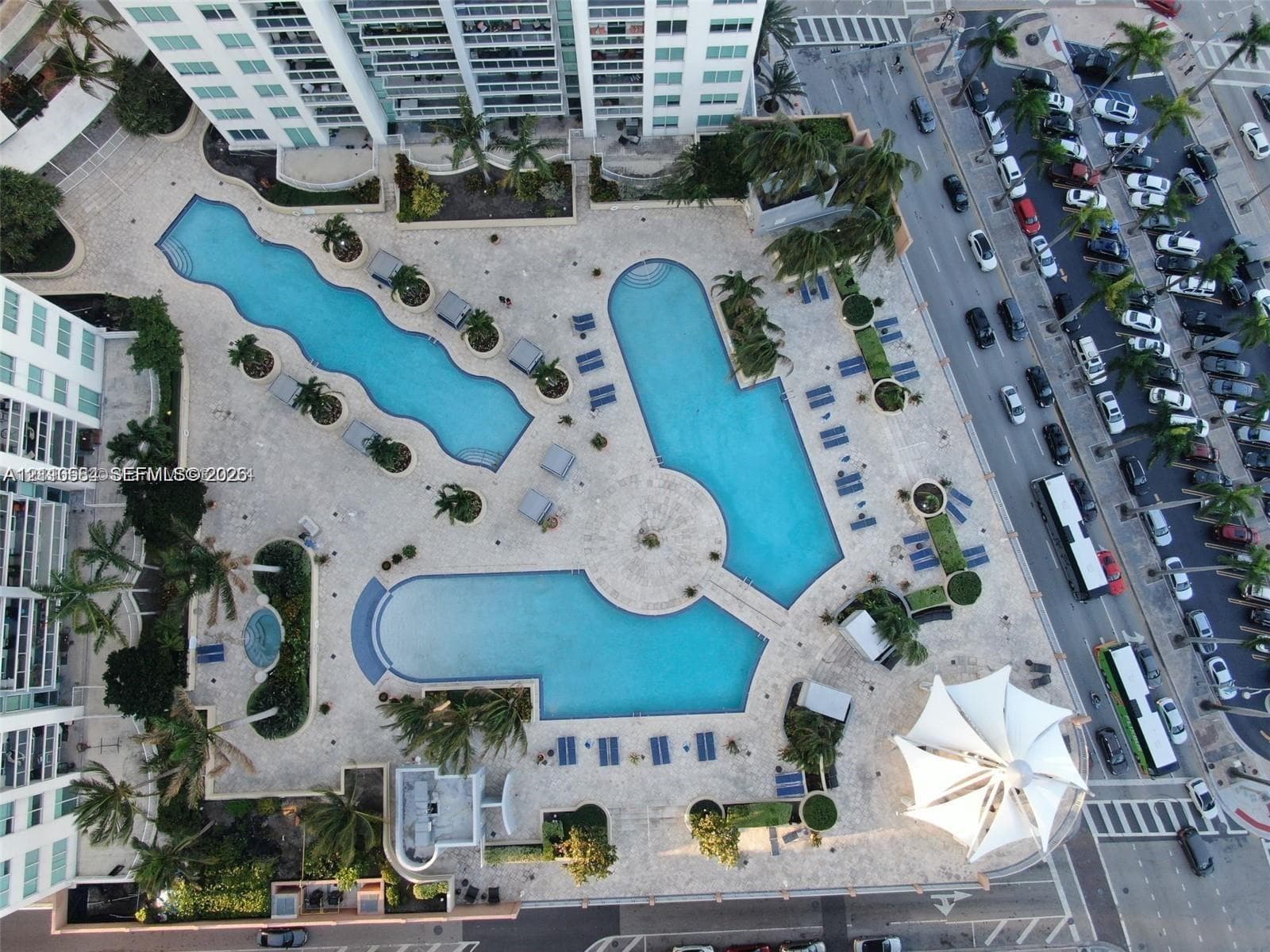 Expansive pool deck featuring multiple swimming pools, stone-paver hardscaping, mature palm trees, and a tension-membrane canopy structure