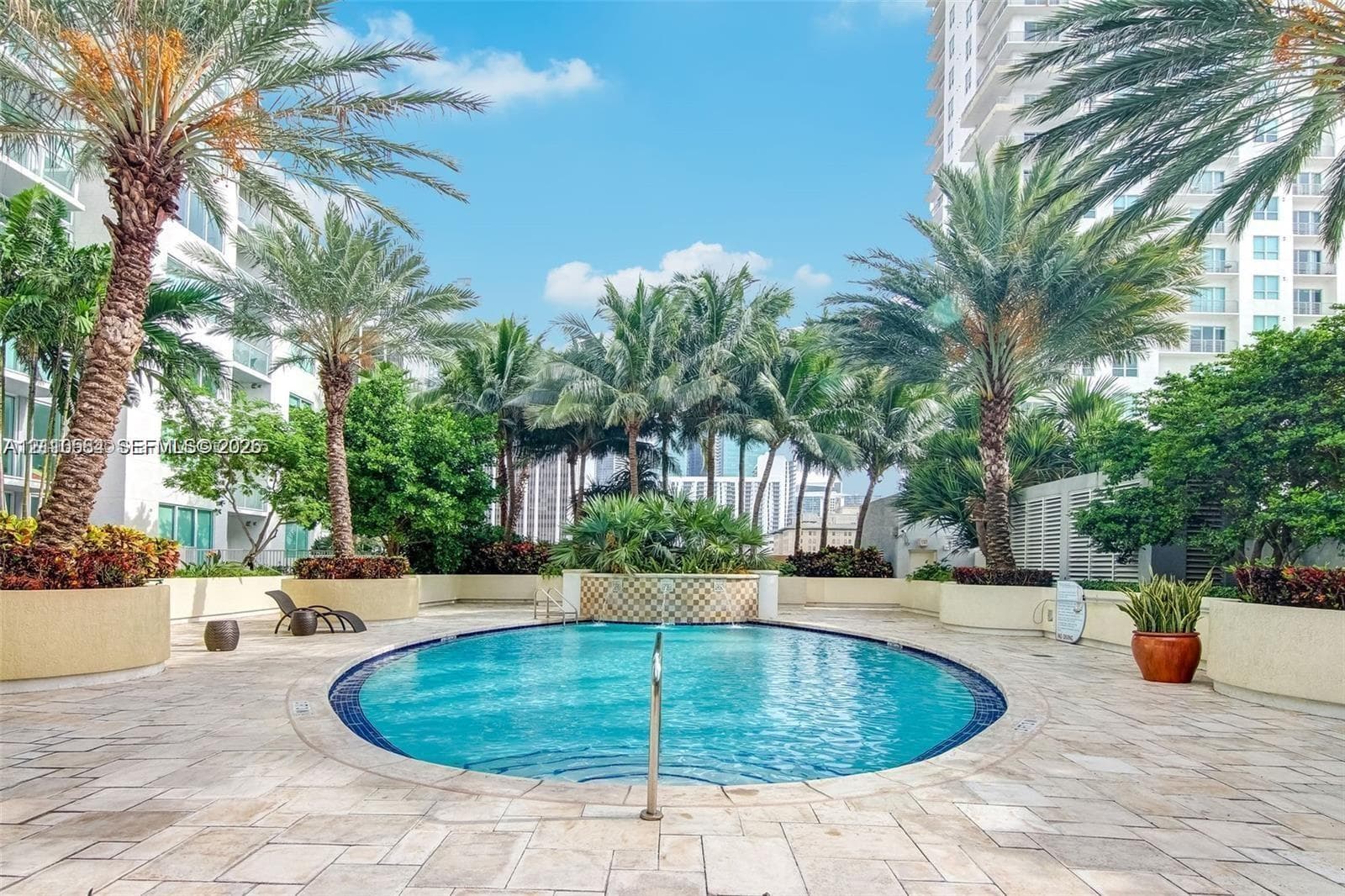 Oval swimming pool featuring natural stone paving, integrated planter beds, and mature palm trees