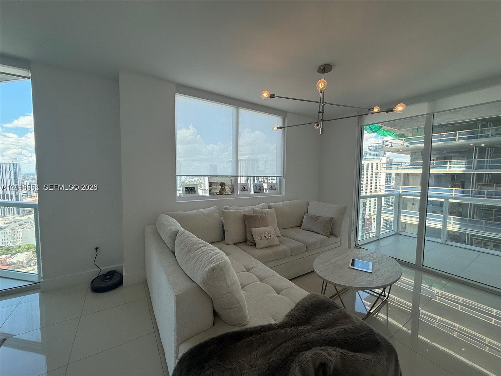 Open-concept interior featuring large-format tile flooring, floor-to-ceiling glass windows, a modern multi-arm chandelier, and sliding glass doors leading to a balcony