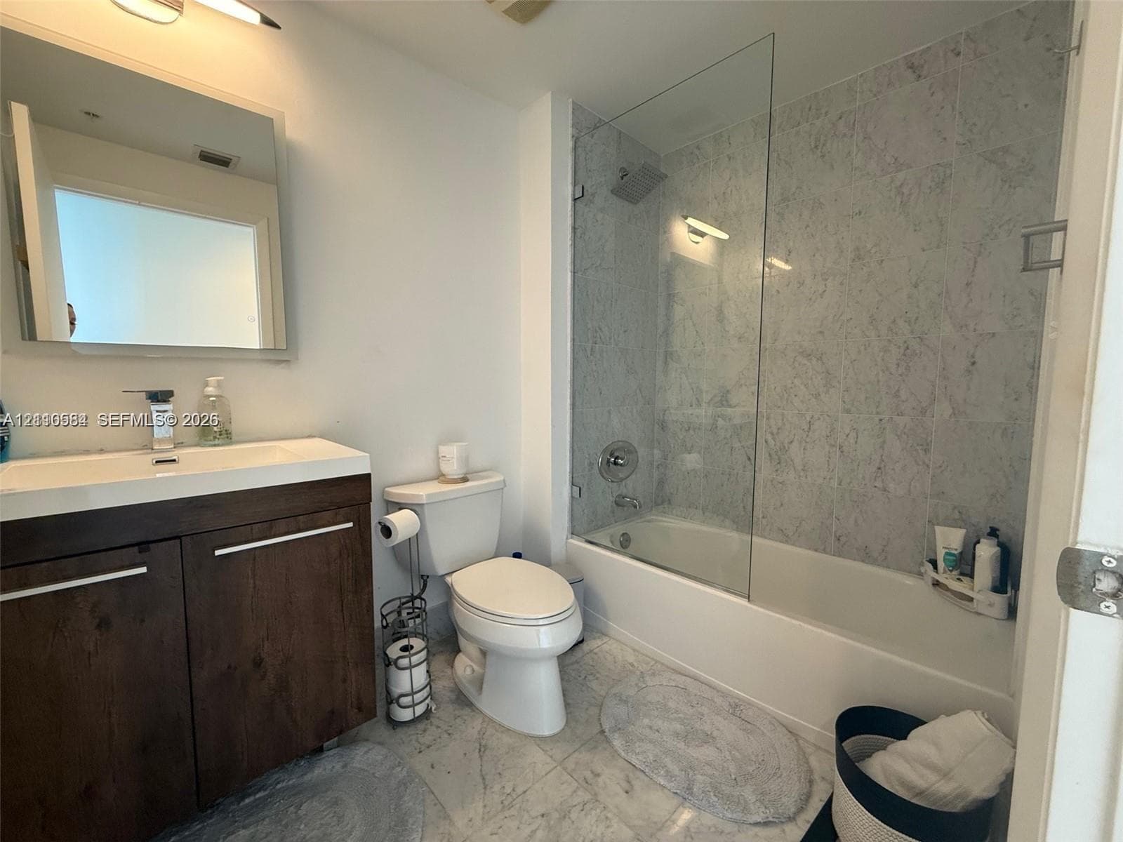 Bathroom featuring a dark wood-finish vanity with a rectangular sink, stone-surround tub with a glass panel, and light-toned tile flooring