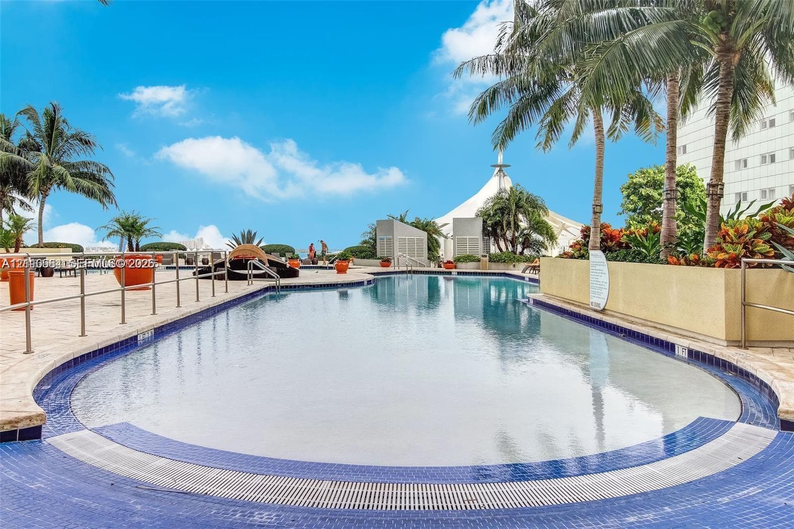 In-ground swimming pool with tile surround, stone-paved deck, metallic perimeter railing, and integrated landscaping with mature palm trees