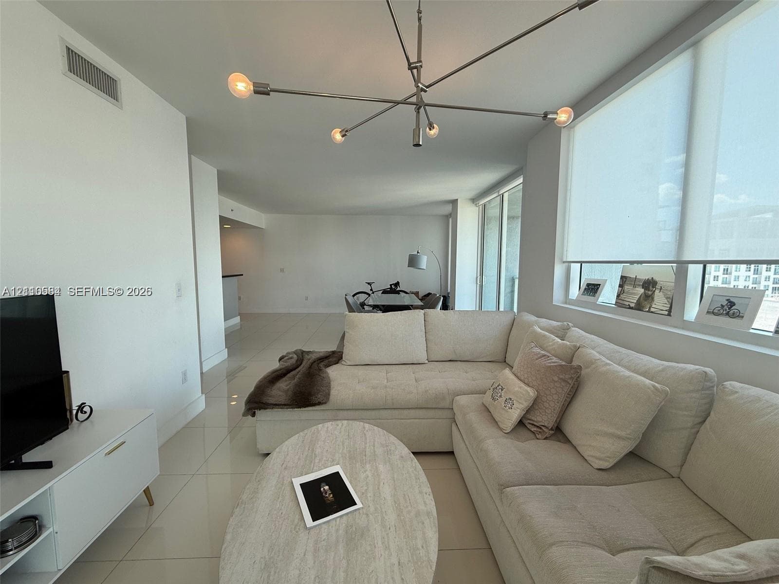Open-concept space featuring floor-to-ceiling windows with roller shades, large-format tile flooring, and a multi-arm contemporary chandelier
